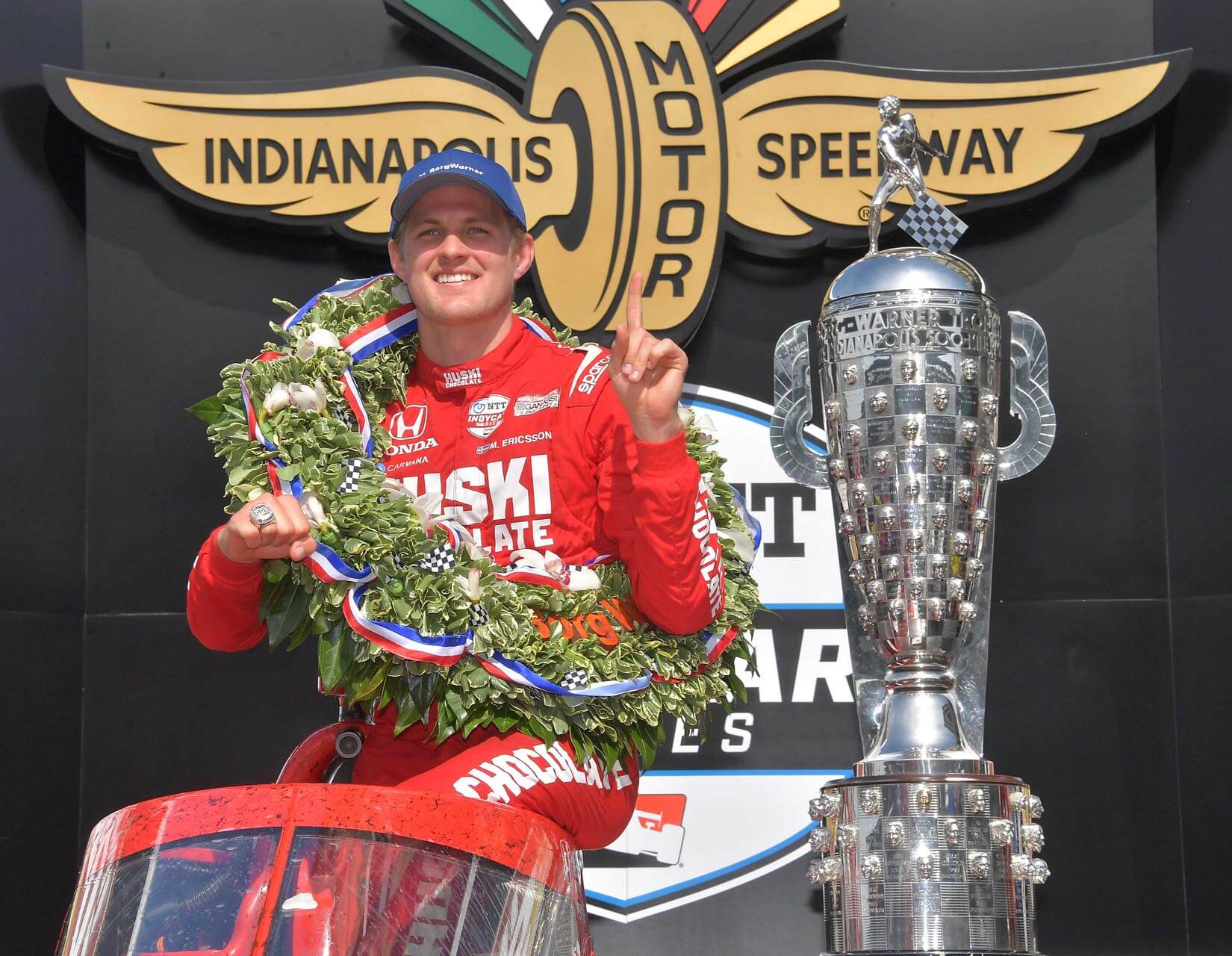 a person in a red race car with a wreath around his neck and a trophy