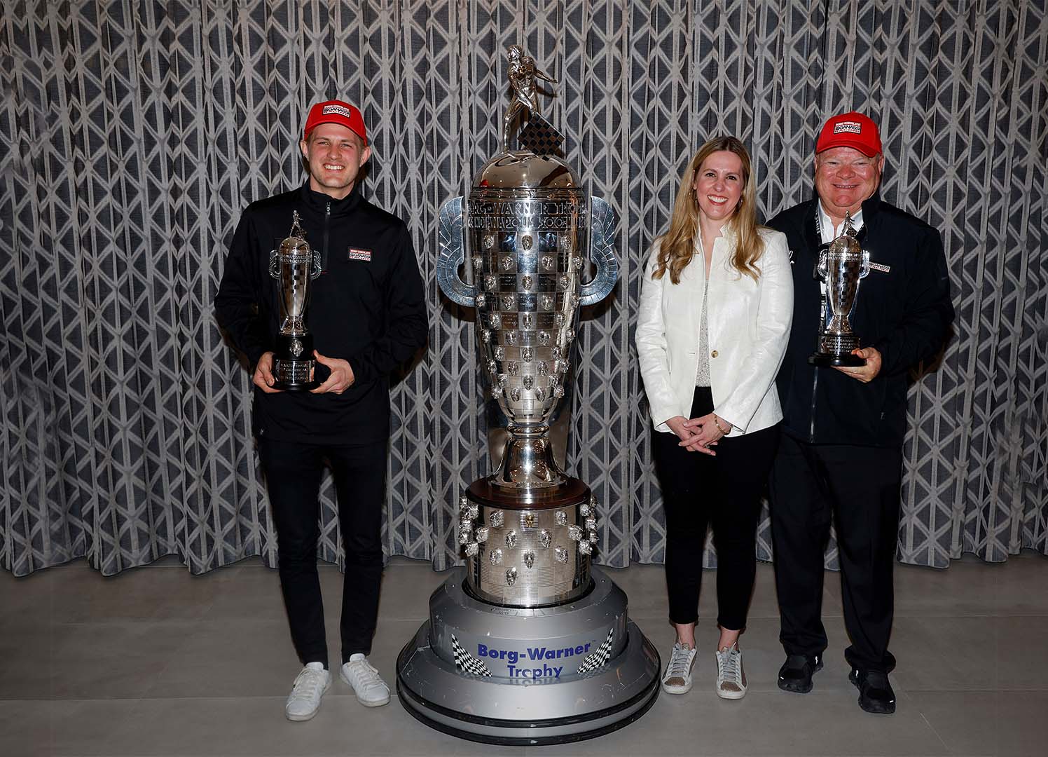 Three people in line stand around large silver trophy holding smaller trophies and smiling