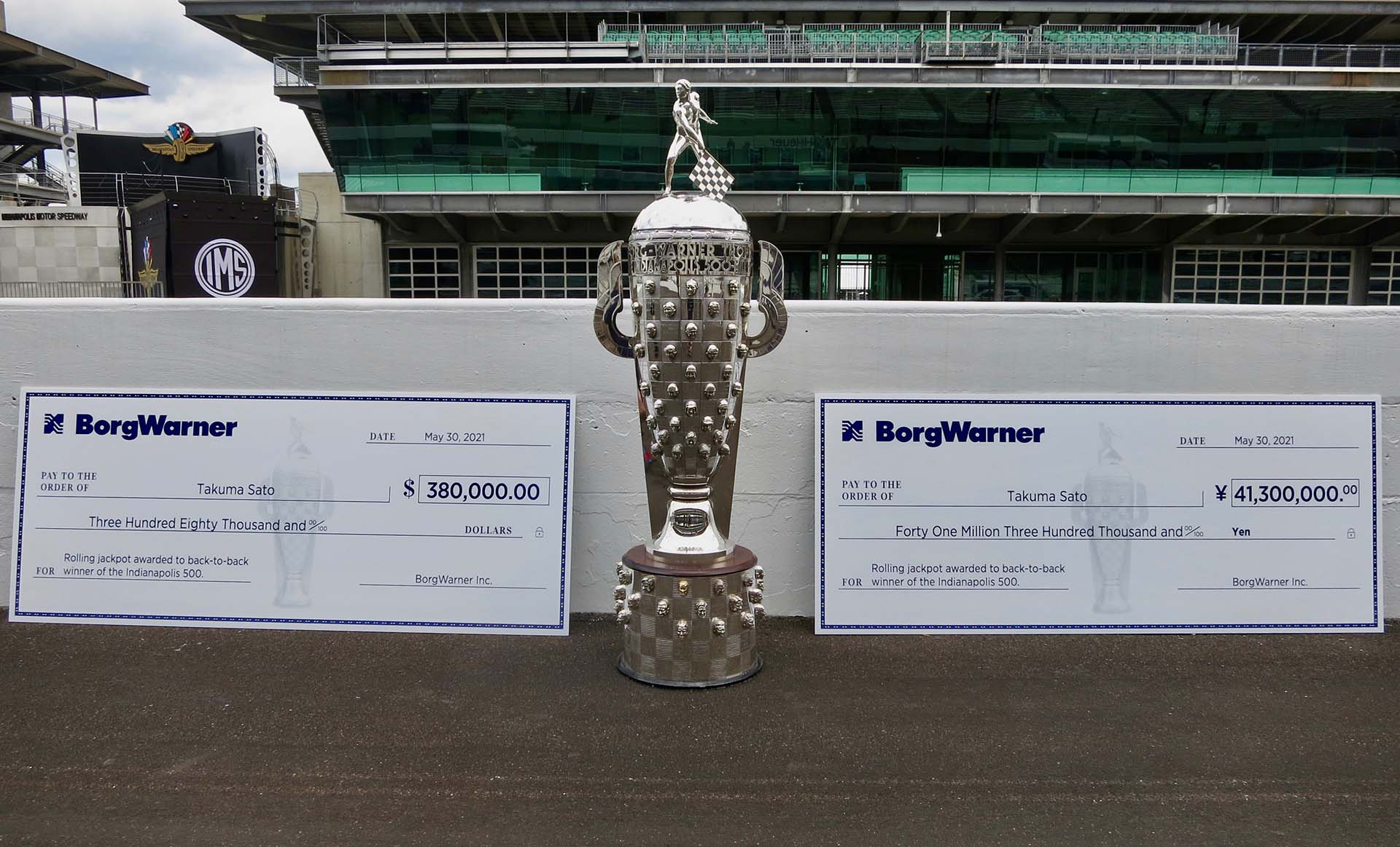 Large silver trophy with two large checks on either side on race track with stands in background