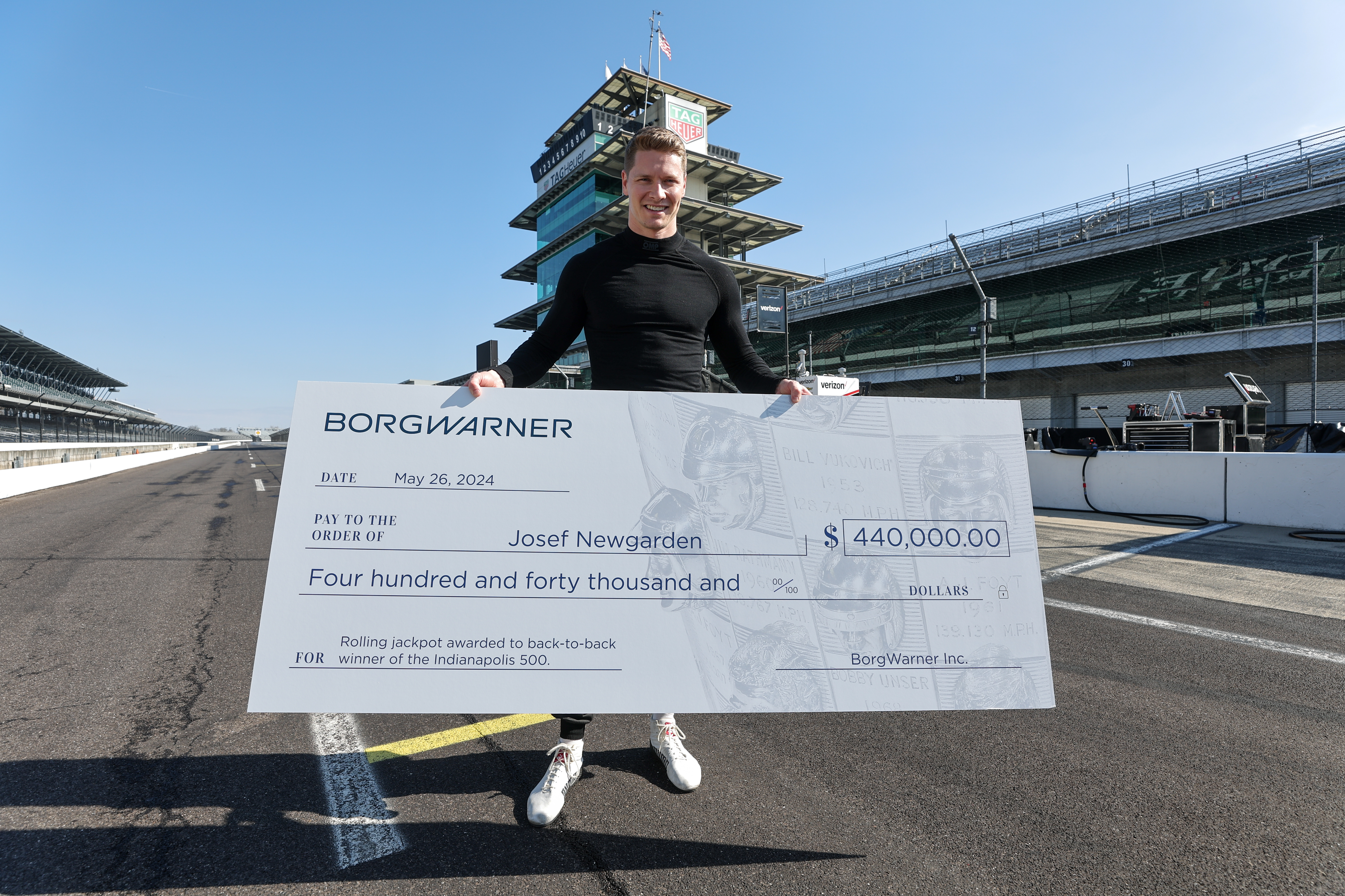 Man in black shirt stands on speedway track holding large check for $440,000