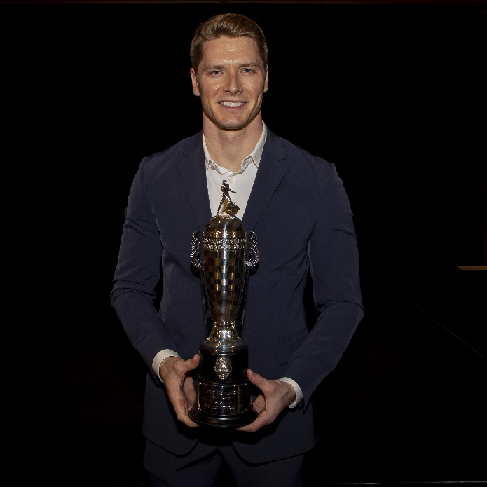 Man in suit jacket smiles and holds trophy